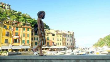 Una mujer caminando con un sombrero y una mochila viajando en Portofino, Italia, una ciudad turística de lujo en Europa. video