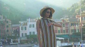 una donna con orologio, cappello, occhiali da sole, abito a righe in viaggio a portofino, in italia, una località turistica di lusso in europa. video