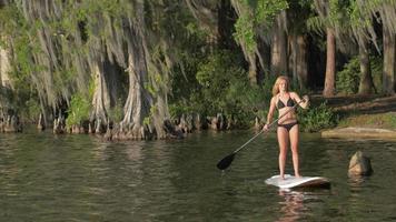 una giovane donna sup stand-up paddleboarding su un lago circondato da alberi.