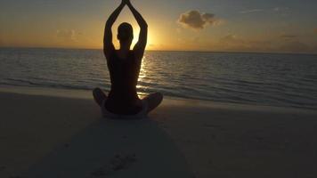 une femme fait une pose de yoga assise sur la plage avec les mains au coucher du soleil.