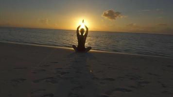 A woman does a seated yoga pose on the beach with hands up at sunset.