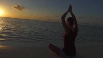 Eine Frau macht eine sitzende Yoga-Pose am Strand mit erhobenen Händen bei Sonnenuntergang.