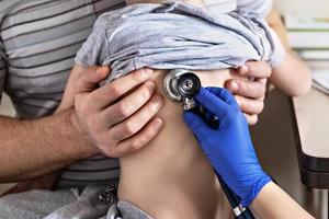 Little girl in the arms of her father in the doctor's office at the clinic. The doctor examines the child, listens to the lungs with a phonendoscope. Treatment and prevention of respiratory infections. photo