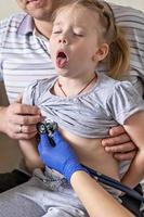 Little girl in the arms of her father in the doctor's office at the clinic. The doctor examines the child, listens to the lungs with a phonendoscope. Treatment and prevention of respiratory infections. photo