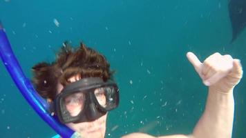 A man takes a selfie while snorkeling with manta rays swimming behind him.