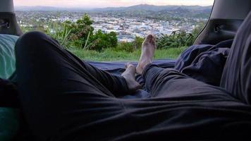 POV of legs and feet of man lying down camping in the back of his suv car vehicle above a city. video