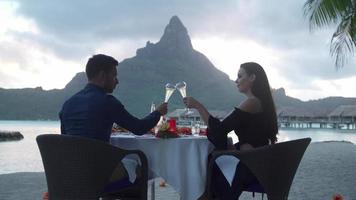 um casal de homem e mulher brindando com champanhe na praia de um resort de ilha tropical ao pôr do sol em bora bora.