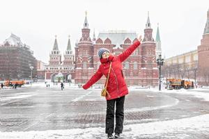 una hermosa joven con una chaqueta roja camina por la plaza manezhnaya en moscú durante una nevada y una ventisca. los quitanieves están trabajando en segundo plano. foto