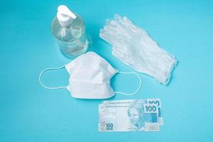 Recipiente con gel de alcohol, guantes, mascarilla quirúrgica y dinero real brasileño, sobre el fondo azul claro