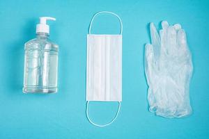 Container with alcohol gel, gloves and surgical mask on the light blue background