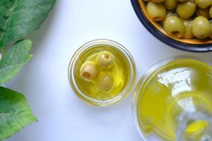 Botella de aceite de oliva y aceitunas frescas en un recipiente sobre fondo blanco. foto