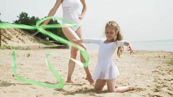 Mother and daughter in white bathing suits dancing with a gymnastic ribbon on a sandy beach. video