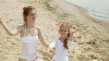 mãe e filha em maiôs brancos, dançando com uma fita de ginástica em uma praia arenosa.