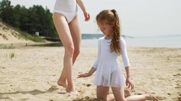 madre e hija en trajes de baño blancos bailando con una cinta de gimnasia en una playa de arena.