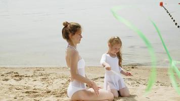 madre e hija en trajes de baño blancos bailando con una cinta de gimnasia en una playa de arena. amanecer de verano