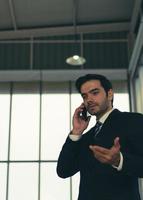 Candid portrait of young handsome caucasian confident businessman in black suit with tie, using smartphone to talk with client in office photo