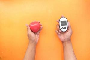 Mano sujetando el medidor de glucosa y el símbolo en forma de corazón sobre fondo naranja foto
