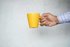 Mano de hombre sosteniendo una taza de color amarillo contra la pared blanca foto