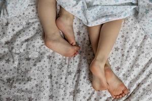 Close-up of two pairs of toddler girls feet on the bed under the blanket