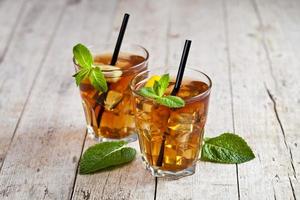 Cold iced tea with lemon, mint leaves and ice cubes in two glasses on rustic wooden table. photo