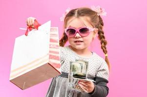 niña con gafas con un paquete y dinero sobre un fondo rosa, niño y compras. foto