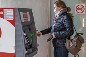 A woman wearing a protective medical mask on her face inserts a credit bank card from an ATM photo