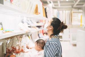 madre asiática con una máscara sostiene a su bebé de compras en una tienda por departamentos. foto