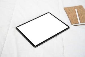 Top view of tablet with a white screen lies on a bed with notebook and digital pen.