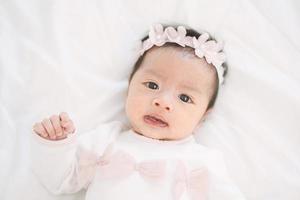 Adorable little baby girl in a beautiful dress with flower band lying on a white blanket.