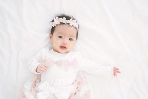 Adorable little baby girl in a beautiful dress with flower band lying on a white blanket.