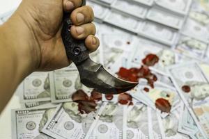 Mano sujetando un cuchillo karambit en la pila de paquetes de billetes de 100 dólares estadounidenses ensangrentados foto