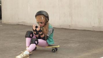 Fröhliches kleines Mädchen sitzt auf einem gelben Skateboard und surft mit dem Handy im Internet video
