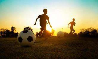 silueta deporte de acción al aire libre de un grupo de niños que se divierten jugando fútbol soccer para hacer ejercicio en la zona rural de la comunidad bajo el crepúsculo del cielo del atardecer.