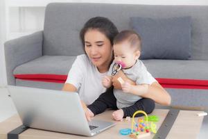 Young asian mother working on laptop computer while care daughter girl at home, mom and baby girl sitting using notebook video call, parent and bonding, indoors, family and business concept. photo