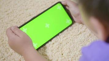 niño asiático preescolar con gadget jugando videojuegos digitales en el teléfono móvil en casa. niño pequeño que usa y sostiene una pantalla verde de teléfono inteligente en la mano, concepto de generación de tecnología video