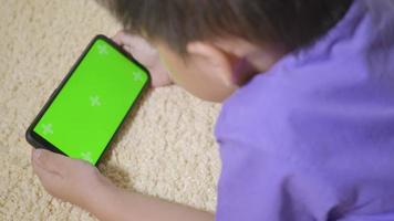 asiatisk pojke förskola med prylar som spelar videospel digitalt på mobiltelefonen hemma. litet barn använder och håller en smartphone grön skärm i handen, teknik generation koncept