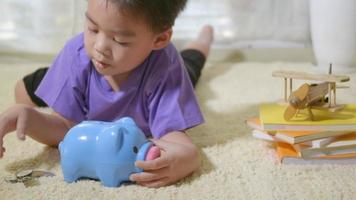 Happy Asian kid boy preschool putting pin money coins into blue faced piglet slot. Little child putting coin into piggy bank for saving with pile of coins at home, Investment education concept video