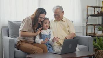 Happy family senior grandfather, daughter and granddaughter talking online via video chat using laptop computer with father, old grandpa embracing little grand child and mother in living room at home