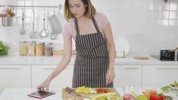 Happy Asian beautiful young woman in kitchen following recipe on digital tablet computer. Female cooking salad online video clip on website via tablet content on internet technology. modern food