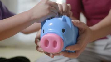 Happy Asian kid boy preschool and mother putting pin money coins into piglet. Little child putting coin into piggy bank for saving with pile of coins at home, Investment education concept video