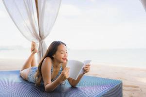 Portrait beautiful young asian woman reading book with happy smile relax in lounge bed chair on the beach sea ocean for leisure photo