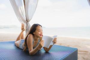 Portrait beautiful young asian woman reading book with happy smile relax in lounge bed chair on the beach sea ocean for leisure photo