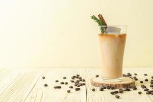 Pouring milk in black coffee glass with ice cubes, cinnamon, and rosemary on wood background photo