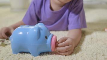 Happy Asian kid boy preschool putting pin money coins into blue faced piglet slot. Little child putting coin into piggy bank for saving with pile of coins at home, Investment education, Slow motion video