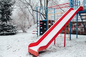 Snow covered baby swing in winter - empty playground - red plastic swing chair in the cold