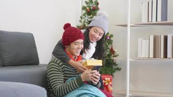 Happy Asian beautiful young family couple husband and wife in home living room, woman hide surprise giving gift box her boyfriend present during play smartphone, winter holidays and valentine concept video