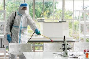 Worker from decontamination services wearing personal protective equipment or ppe including suit, face shield and mask. He uses disinfectant to spray and clean scientist lab photo