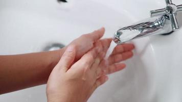 Primer plano de las manos de una mujer joven usando jabón y lavándose las manos bajo el grifo de agua, las mujeres se lavan las palmas con burbujas blancas en el lavabo del baño del hotel para protegerla de la covid-19 video