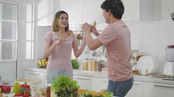 Happy Asian beautiful family couple husband and wife singing in kitchenware microphones in kitchen together having fun dance listen music at home. Two people dancing video