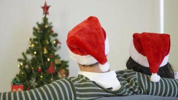 Feliz asiático jovem casal de família, marido e mulher sentados no sofá, usando um chapéu de Papai Noel em frente à lareira no inverno quente de natal em casa, abraçando a sala de estar, vista traseira, câmera lenta video